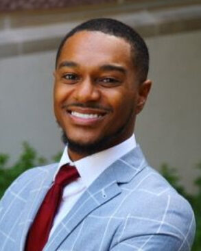 A man in a light blue checkered suit jacket, white shirt, and red tie smiles while standing outdoors in front of greenery and a building.