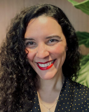 A woman with long curly hair, wearing a black top with gold dots, smiles at the camera. There are green leaves and a wooden wall in the background.