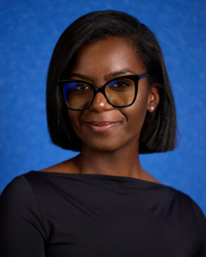A woman with short black hair wearing glasses and a black top poses against a solid blue background.