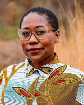 A person with short hair and glasses wearing a patterned shirt stands outdoors, looking at the camera with a neutral expression. Tall grass and blurred foliage are in the background.