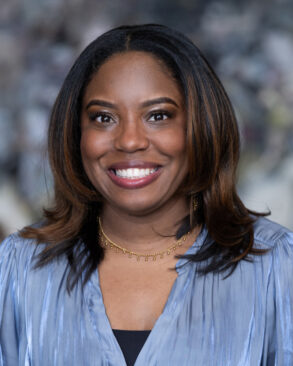 A woman with medium-length dark hair, wearing a light blue blouse and a gold necklace, smiles at the camera in front of a blurred background.