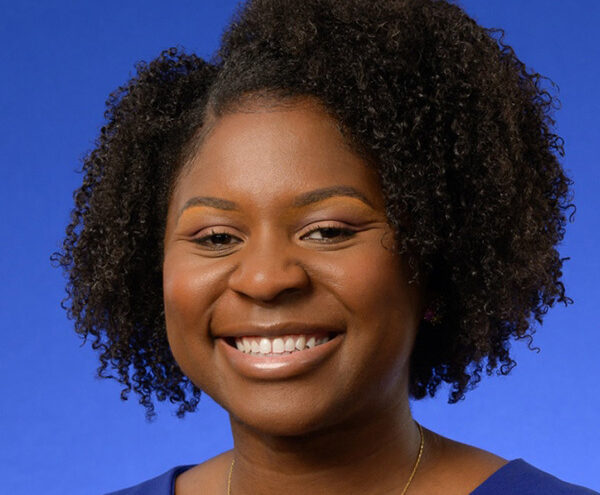 A woman with curly black hair, wearing a blue top, smiles at the camera against a solid blue background.
