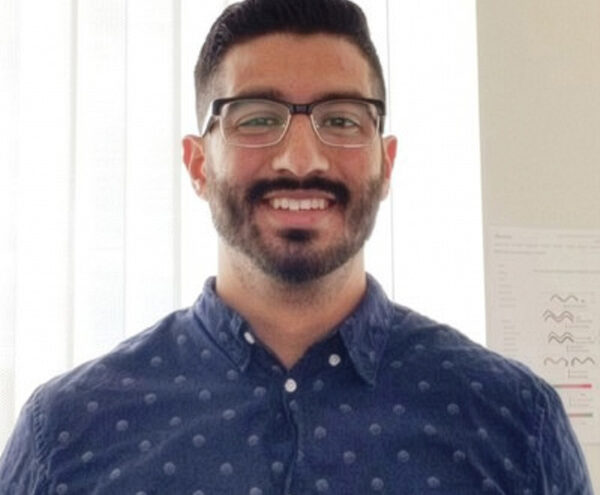 A man with glasses, dark hair, and a beard is smiling while standing indoors in front of a window with vertical blinds. He is wearing a blue, patterned button-up shirt.