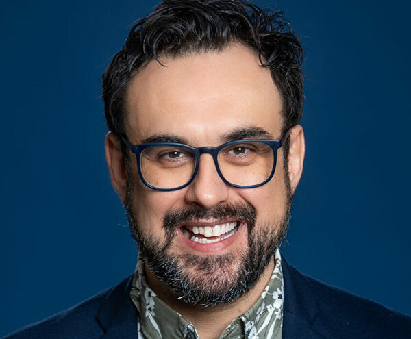 A man with dark hair, beard, and glasses is smiling at the camera. He is wearing a dark blazer over a patterned shirt against a blue background.