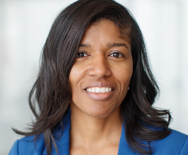 Woman with medium-length dark hair wearing a blue blazer, smiling at the camera with a neutral, blurred background.