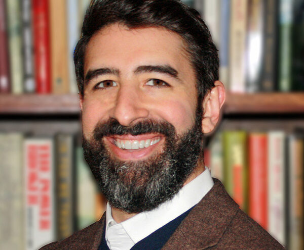 A man with dark hair and a beard, wearing a brown blazer and white shirt, smiles in front of a blurred bookshelf.