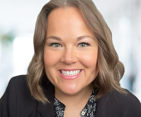 Smiling woman with shoulder-length light brown hair wearing a black blazer and patterned blouse, posed against a blurred indoor background.