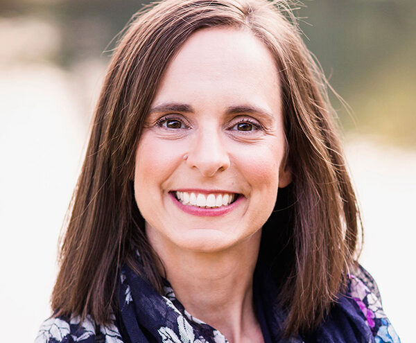 Woman with straight brown hair smiling at the camera, wearing a dark floral scarf and standing outdoors with a blurred background.