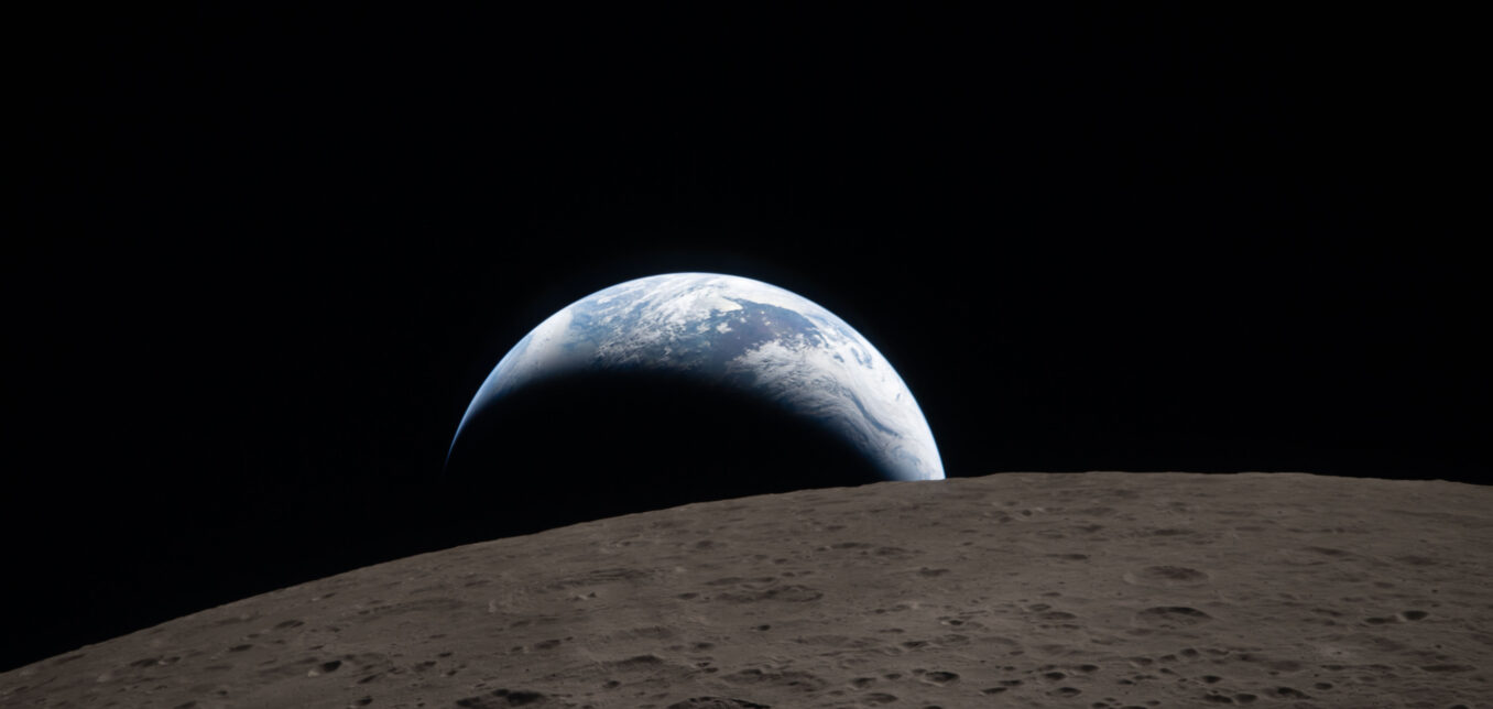 A view of Earth rising over the horizon of the Moon, with the lunar surface in the foreground and the dark space in the background.