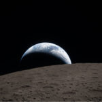 A view of Earth rising over the horizon of the Moon, with the lunar surface in the foreground and the dark space in the background.