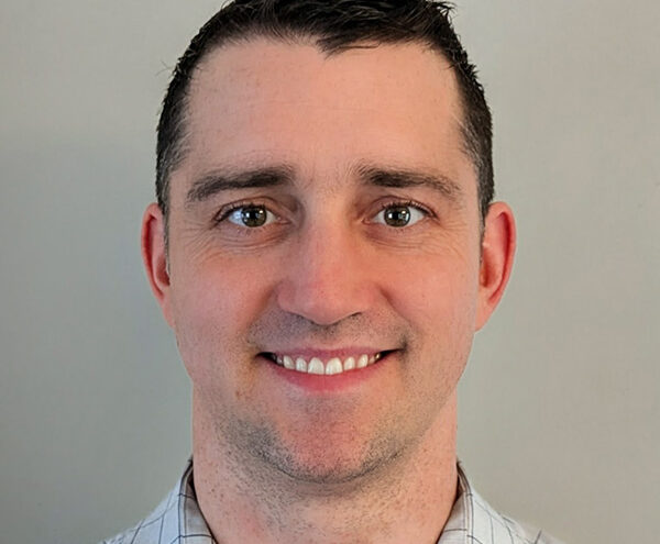 A man with short dark hair and a light complexion, wearing a light-colored checkered shirt, smiling in front of a plain background.