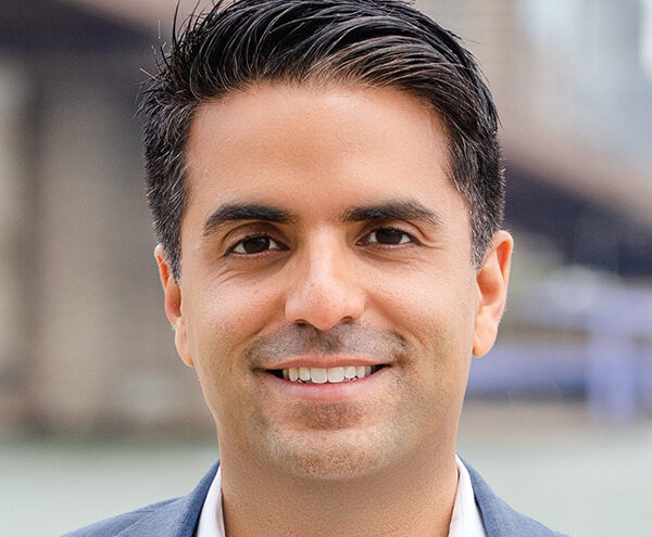 A man with short dark hair, wearing a light blue blazer and white shirt, smiles outdoors with a blurred urban background.