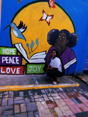 A woman crouches and smiles in front of a colorful mural featuring a book, a hummingbird, a butterfly, a woman, and the words "Hope," "Peace," "Love," and "Joy.