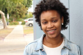A person with short curly hair and a denim jacket smiles while standing outdoors next to a blue wall.