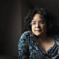 A person with curly dark hair and glasses sits indoors, wearing a patterned top, looking thoughtfully into the distance.
