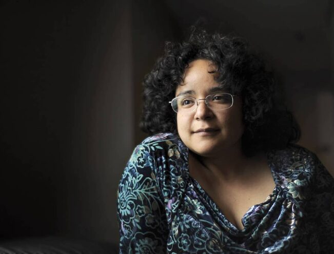 A person with curly dark hair and glasses sits indoors, wearing a patterned top, looking thoughtfully into the distance.