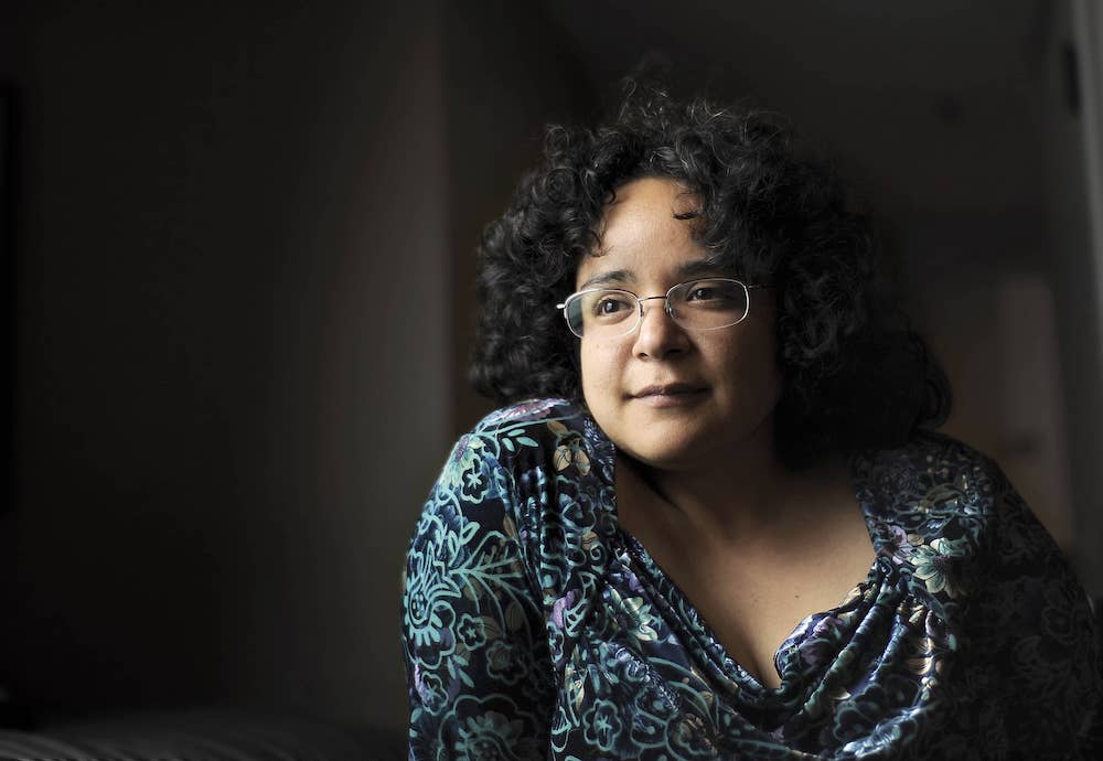 A person with curly dark hair and glasses sits indoors, wearing a patterned top, looking thoughtfully into the distance.