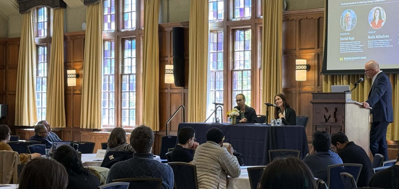 A panel discussion takes place in a wood-paneled room with large windows; two speakers are seated at a table while a moderator stands at a podium. Audience members are seated facing them.