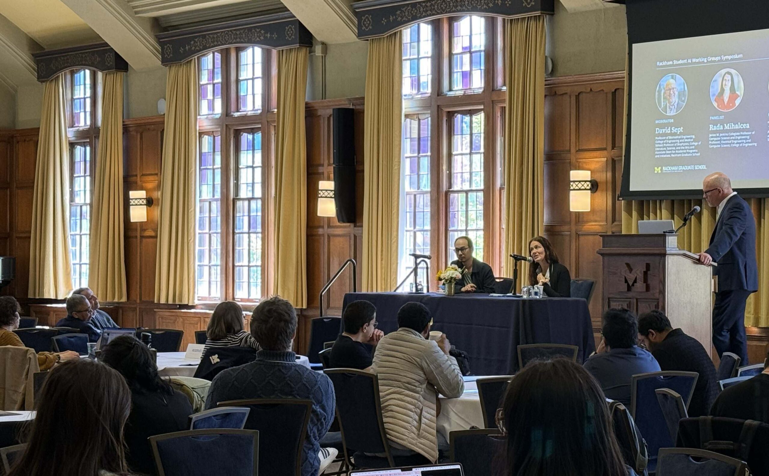 A panel discussion takes place in a wood-paneled room with large windows; two speakers are seated at a table while a moderator stands at a podium. Audience members are seated facing them.
