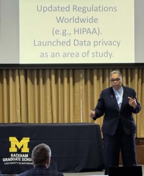 A speaker in a suit presents in front of a screen displaying information about updated regulations and data privacy at Rackham Graduate School, University of Michigan.