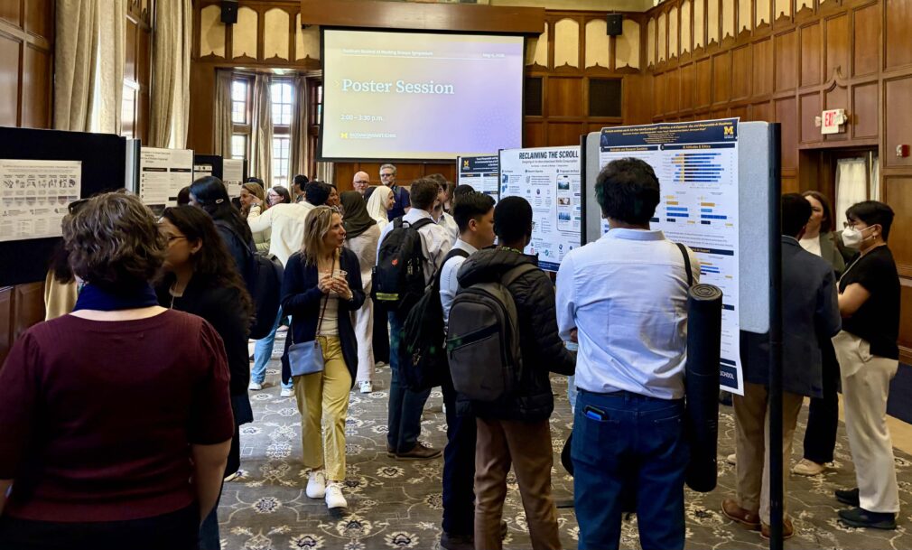People view and discuss research posters at an indoor academic poster session, with a presentation screen visible at the front of the room.