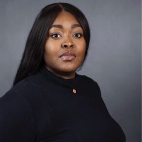 A woman with long dark hair and a black turtleneck poses against a plain gray background, looking directly at the camera with a neutral expression.