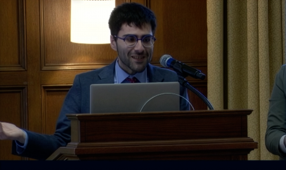 A man in a suit and glasses speaks at a podium with a laptop and microphone, gesturing with one hand in a wood-paneled room.