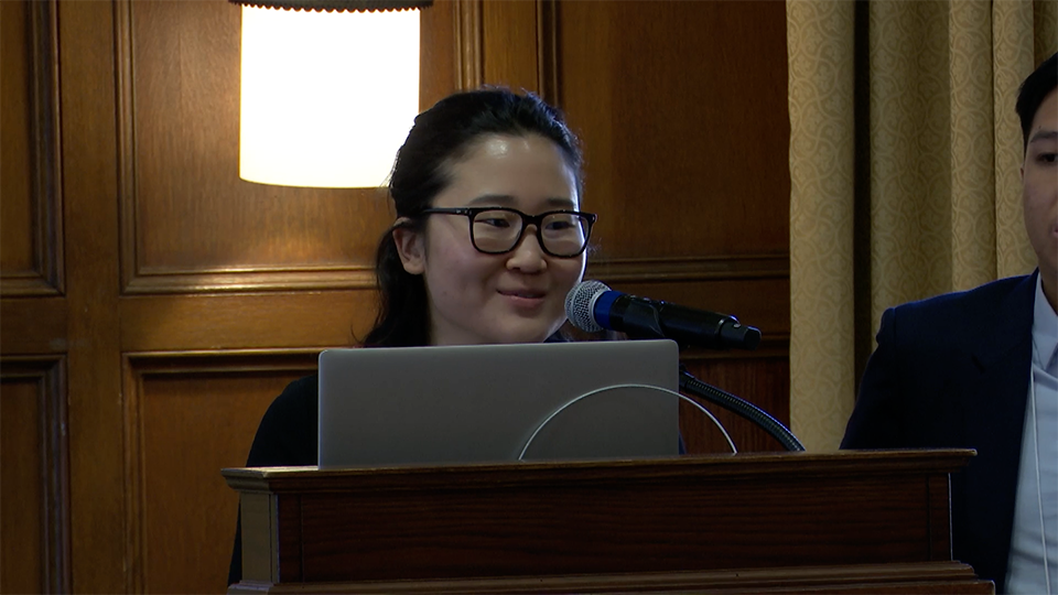 A person with glasses speaks into a microphone at a podium with a laptop, with another person partially visible beside them.