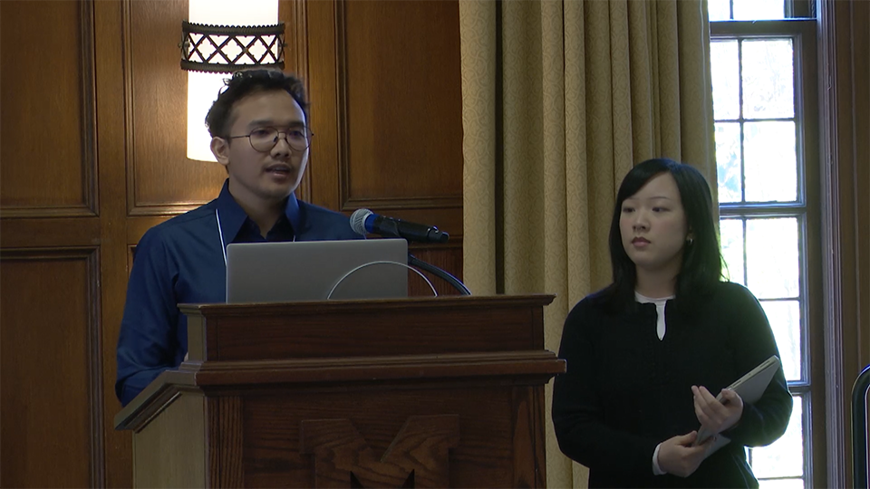 Two people stand at a wooden podium with a laptop and microphone; the person on the left speaks while the person on the right holds a tablet.
