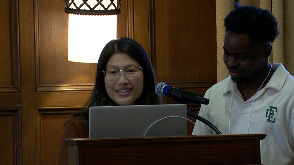 Two people stand behind a podium with a laptop and microphone; one is speaking while the other looks on. A lamp and wood paneling are visible in the background.