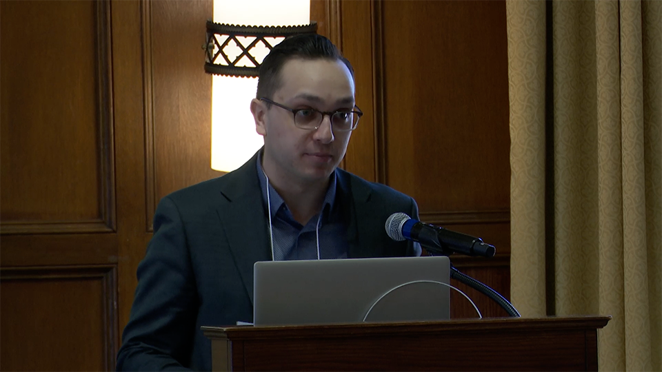 A man in glasses and a suit stands at a podium with a laptop and microphone, speaking in front of wooden paneling and a light fixture.