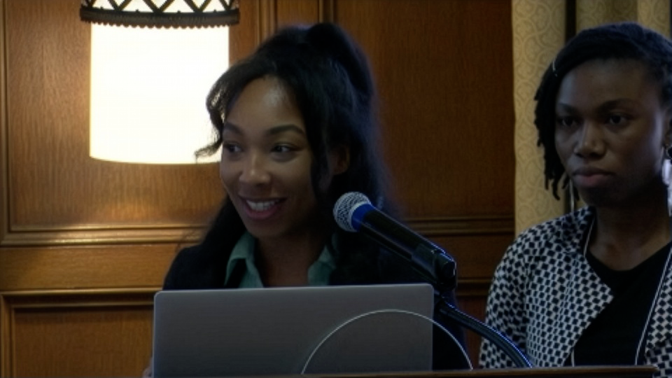 Two women stand behind a podium with a laptop and microphone; one is smiling while speaking, the other looks ahead. A lamp and wood-paneled wall are in the background.