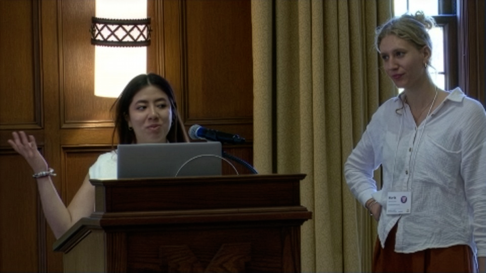 Two women stand at a podium during a presentation; one is speaking at a microphone while the other stands beside her, listening.