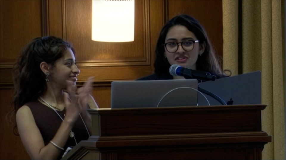 Two women stand at a podium with laptops; one is speaking into a microphone while the other is clapping and looking at her, with wood paneling and a lit lamp in the background.