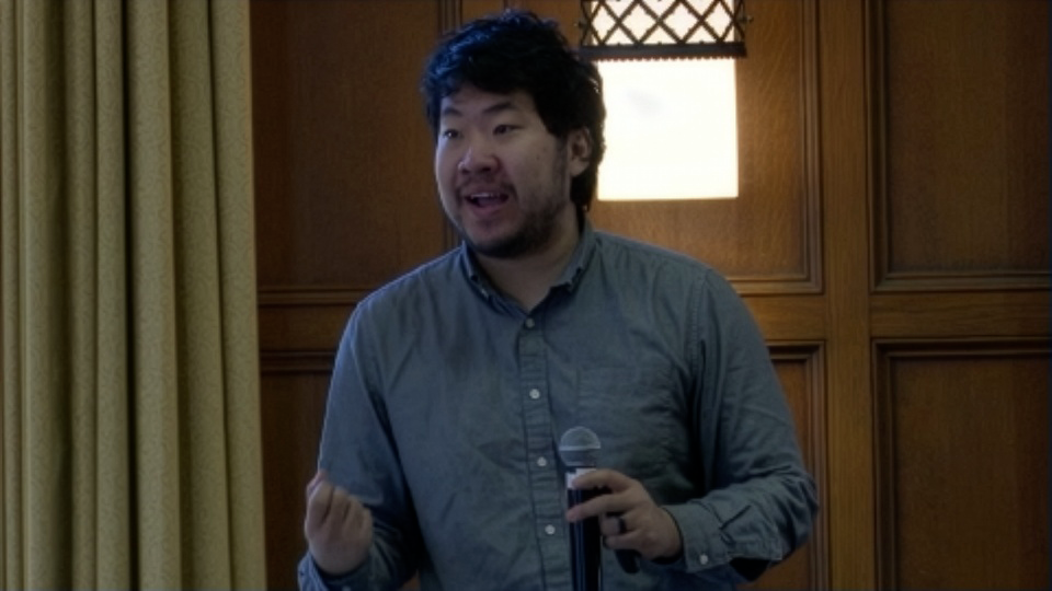 A person in a gray shirt stands indoors holding a microphone and gesturing with one hand, speaking in front of wood-paneled walls and a window with curtains.