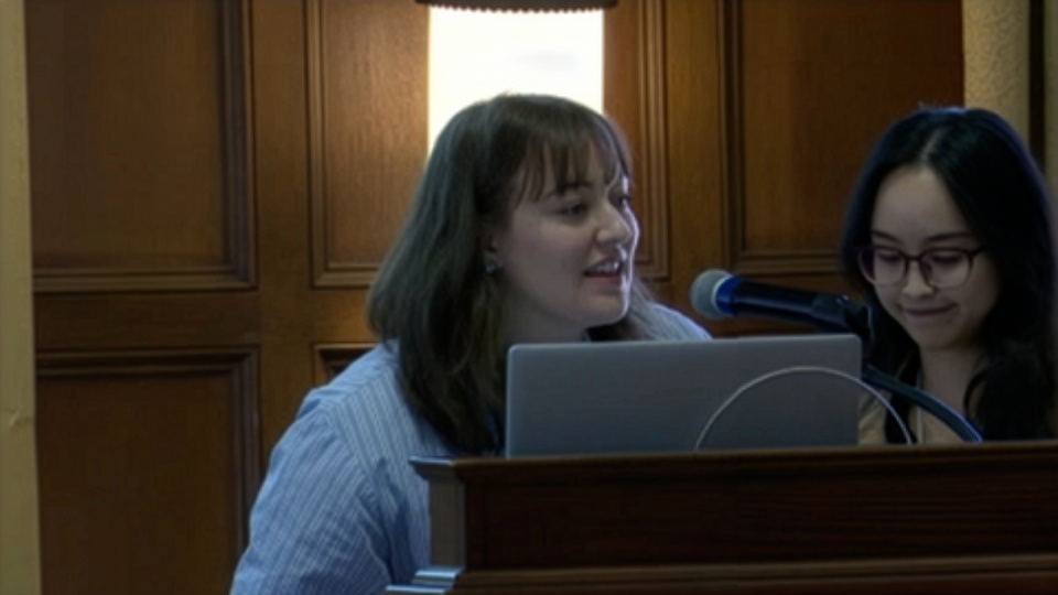 Two women stand at a podium with a laptop and microphone, preparing to speak in a room with wooden paneling in the background.