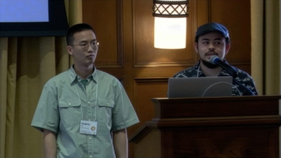 Two people stand at a podium, one speaking into a microphone and the other listening, during a presentation in a wood-paneled room.