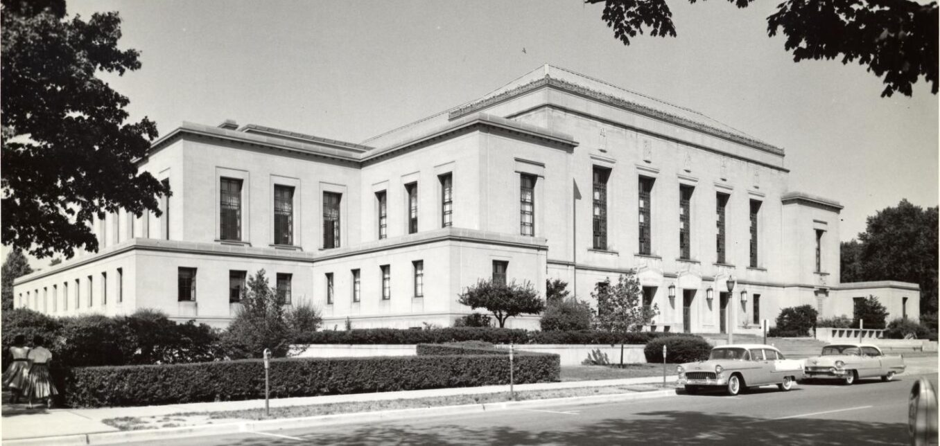 Black and white photo of a large, two-story neoclassical building with columns, surrounded by trees, shrubs, and vintage cars parked along the street in front.