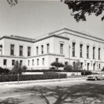 Black and white photo of a large, two-story neoclassical building with columns, surrounded by trees, shrubs, and vintage cars parked along the street in front.