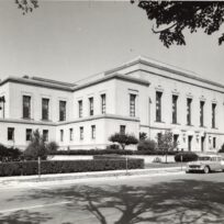 Black and white photo of a large, two-story neoclassical building with columns, surrounded by trees, shrubs, and vintage cars parked along the street in front.