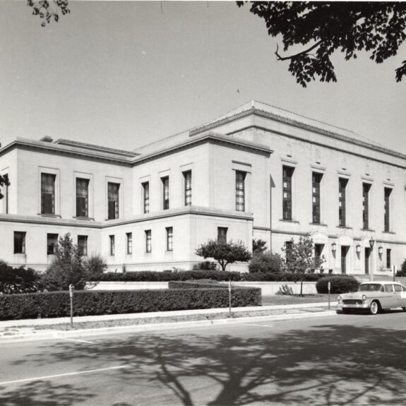 Black and white photo of a large, two-story neoclassical building with columns, surrounded by trees, shrubs, and vintage cars parked along the street in front.