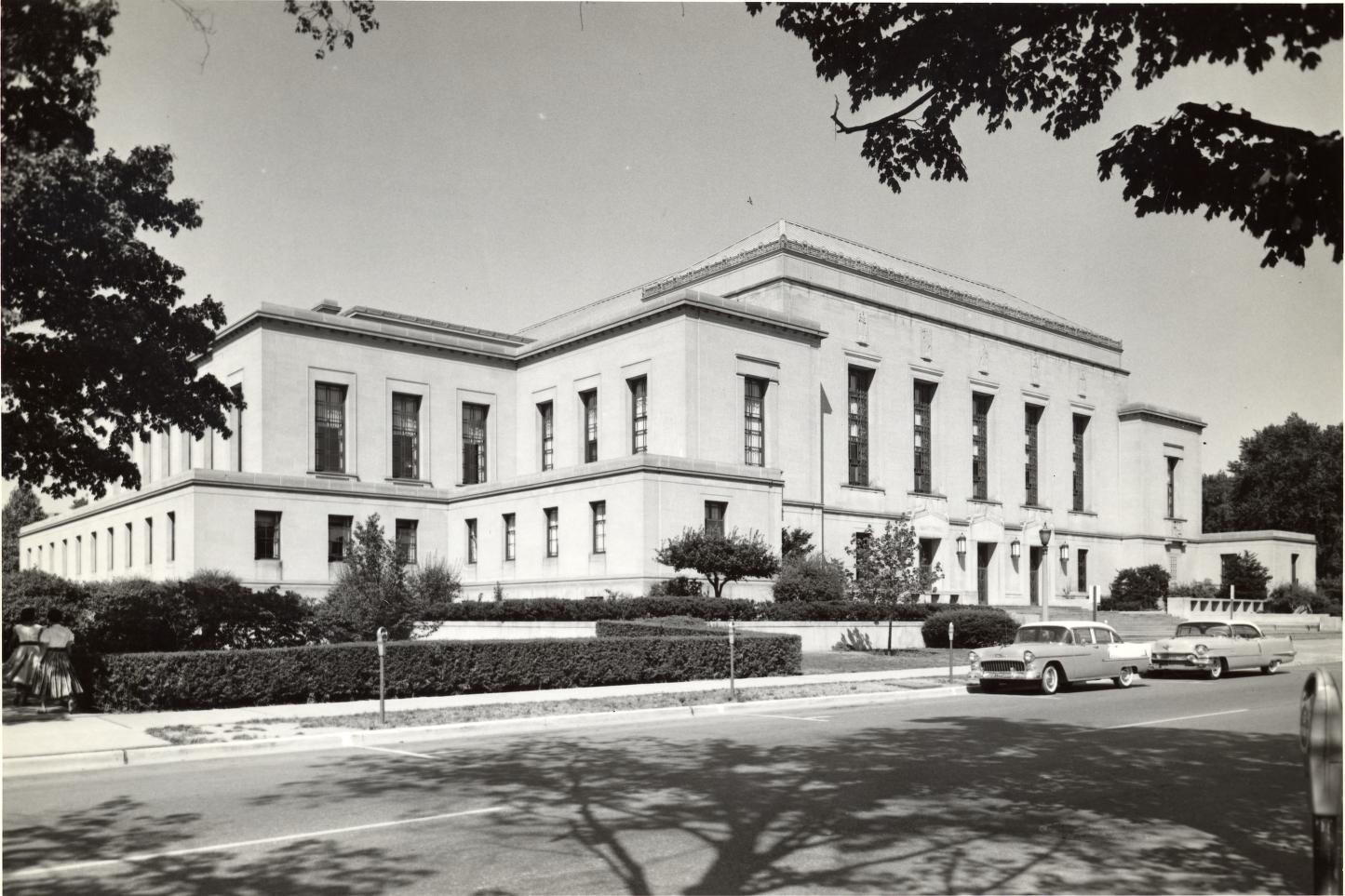 Black and white photo of a large, two-story neoclassical building with columns, surrounded by trees, shrubs, and vintage cars parked along the street in front.
