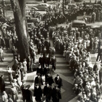 A large crowd stands on either side of a tree-lined street, watching people in dark coats and hats walk in procession along a paved path, with vintage cars parked nearby.
