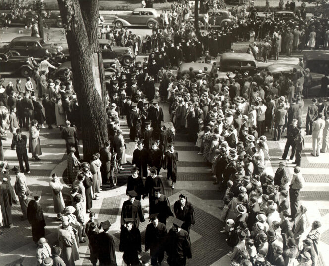 A large crowd stands on either side of a tree-lined street, watching people in dark coats and hats walk in procession along a paved path, with vintage cars parked nearby.
