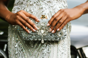 A person in a silver, beaded dress holds a matching embellished clutch bag with both hands, showcasing a detailed and ornate design.