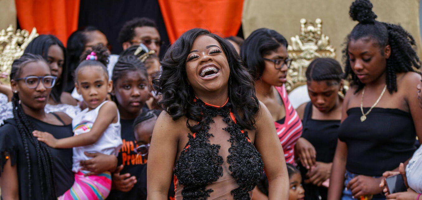 A woman in a black and sheer dress smiles and laughs in front of a group of people, mostly children and women, at an outdoor event.