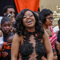 A woman in a black and sheer dress smiles and laughs in front of a group of people, mostly children and women, at an outdoor event.