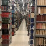 Rows of metal bookshelves filled with books and periodicals line both sides of a well-lit library aisle with a reflective tile floor.