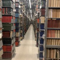 Rows of metal bookshelves filled with books and periodicals line both sides of a well-lit library aisle with a reflective tile floor.