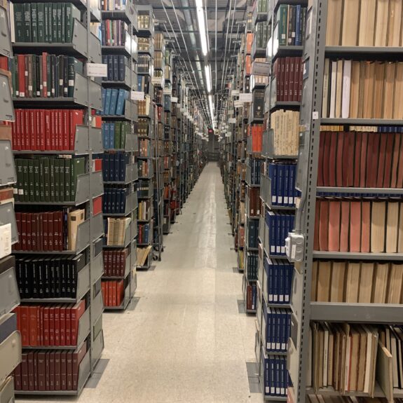 Rows of metal bookshelves filled with books and periodicals line both sides of a well-lit library aisle with a reflective tile floor.